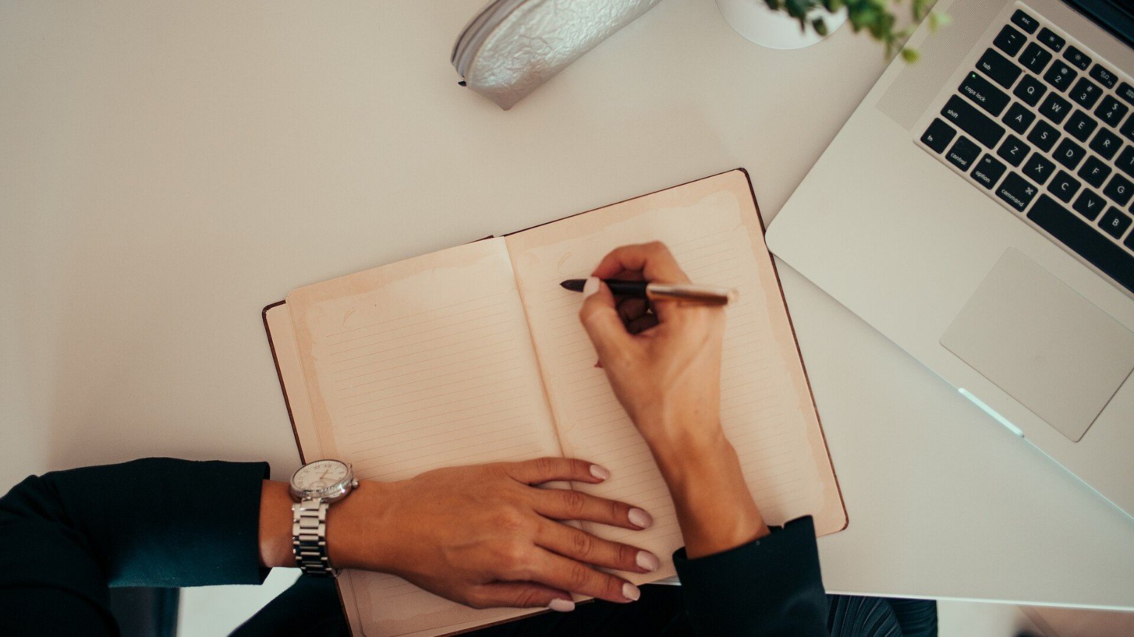 A person writing in a notebook beside a laptop at a workspace.